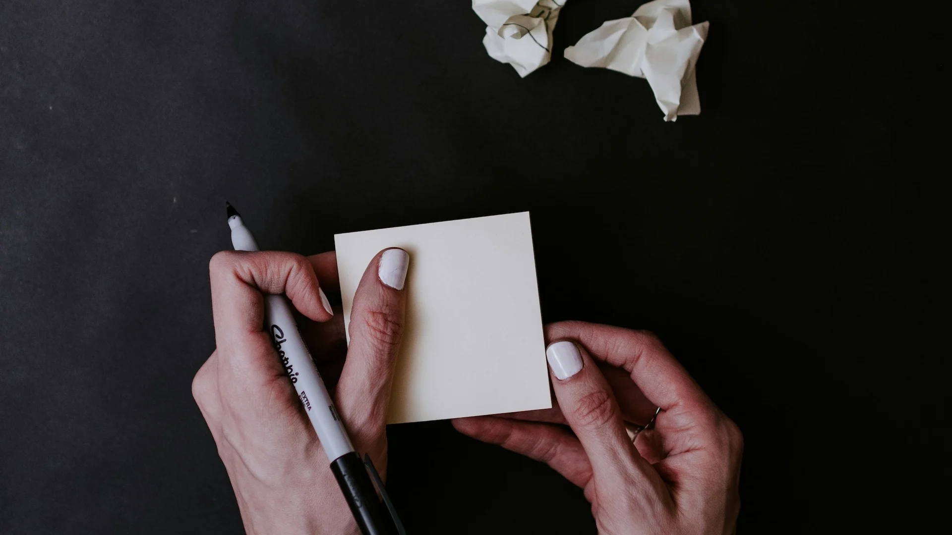 Hands holding a blank note and marker on a dark desk scattered with crumpled paper.